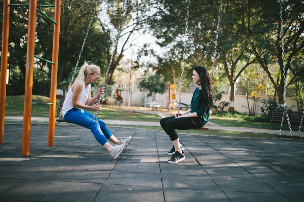 Two young women smile as they sit on playground swigs to where they are facing each other. They're both smiling wide.