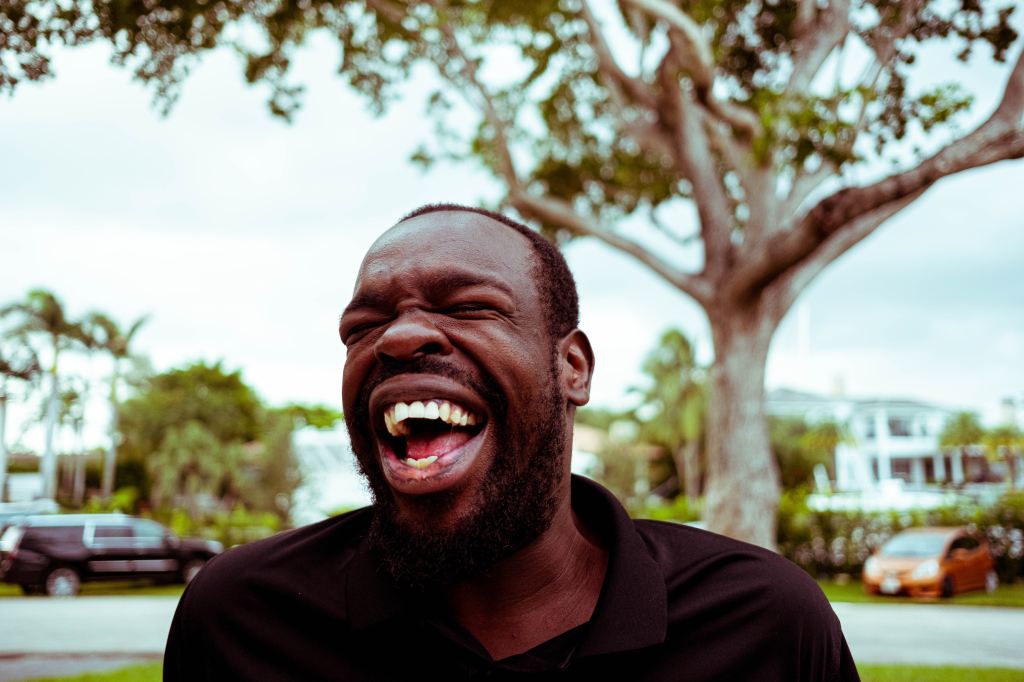 Close up of a man laughing hard. His eyes are closed and his mouth is wide open. He's standing outside.