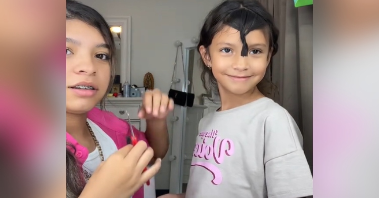 A young girl looks at her younger sister after she attempted to cut her bangs. The little sister smiles, hair still in a twist after getting cut.