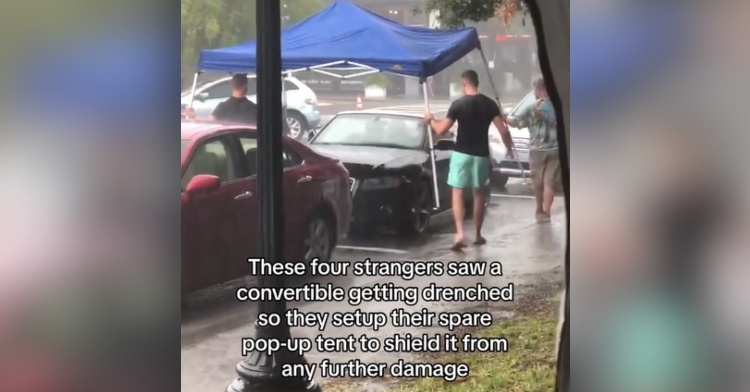 Four people carry a pop-up tent over to a convertible to protect it from the rain.