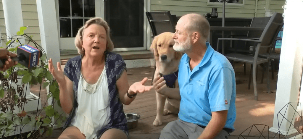 Kebby Kelley and a man sit on their patio steps, talking to a news station. Their dog, Ehmi, sits on the porch just behind them.