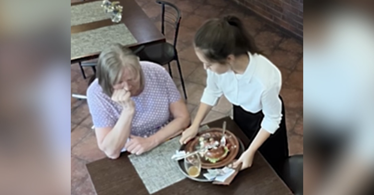 An older woman sits at a restauraunt table looking distraught as a waitress takes away the food sat out in front of her.