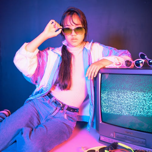 a woman wearing a windbreaker and sunglasses, leaning on a TV