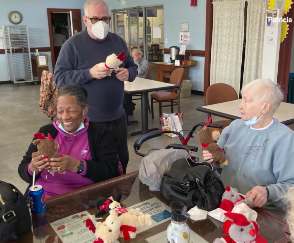 Three elderly people in nursing homes admire the teddy bears they've been given. Two women are sitting, one smiling wide. A man stands nearby, teddy bear in hand.