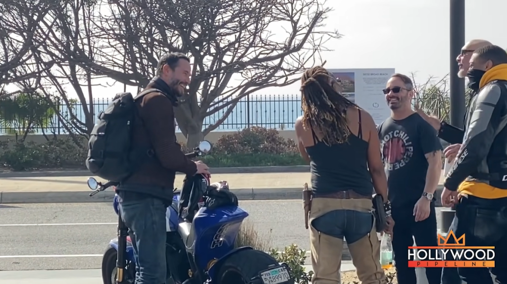 Keanu Reeves smiles wide as he stands next to his motorcycle and chats with four fans.