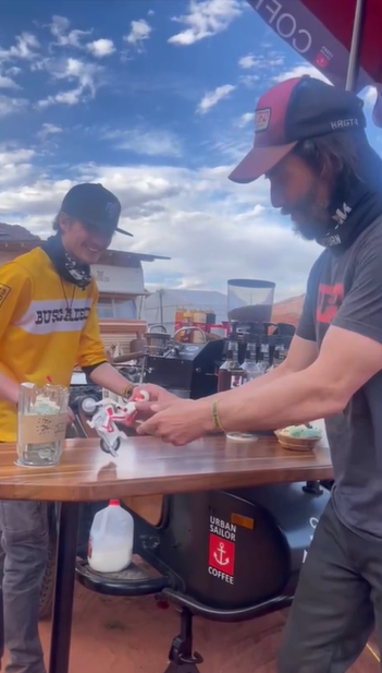 Keanu Reeves plays with a toy of his "Toy Story 4" character, Duke Caboom. Duke is on a motorcycle, so Reeves is making him drive it. An Urban Sailor Coffee employee stands behind the counter, smiling wide at the toy.