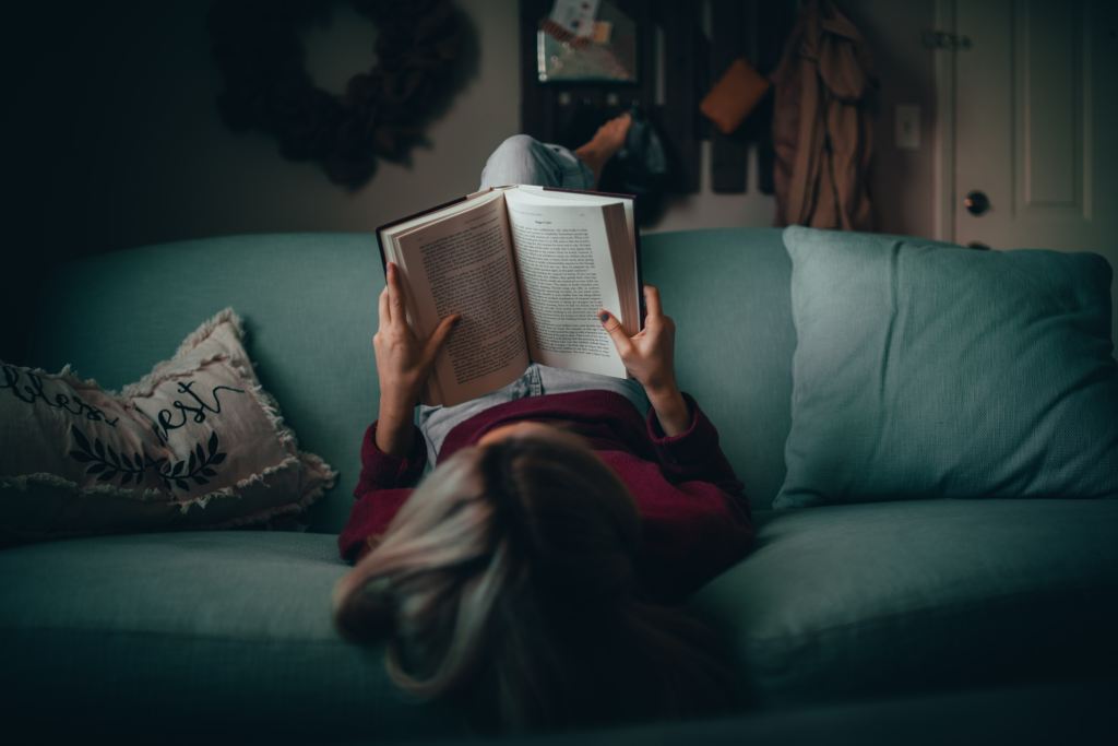 Someone sits on a couch with their back on the seat and their legs dangling over the back. They're reading a book. From our view we see the inside of the pages.