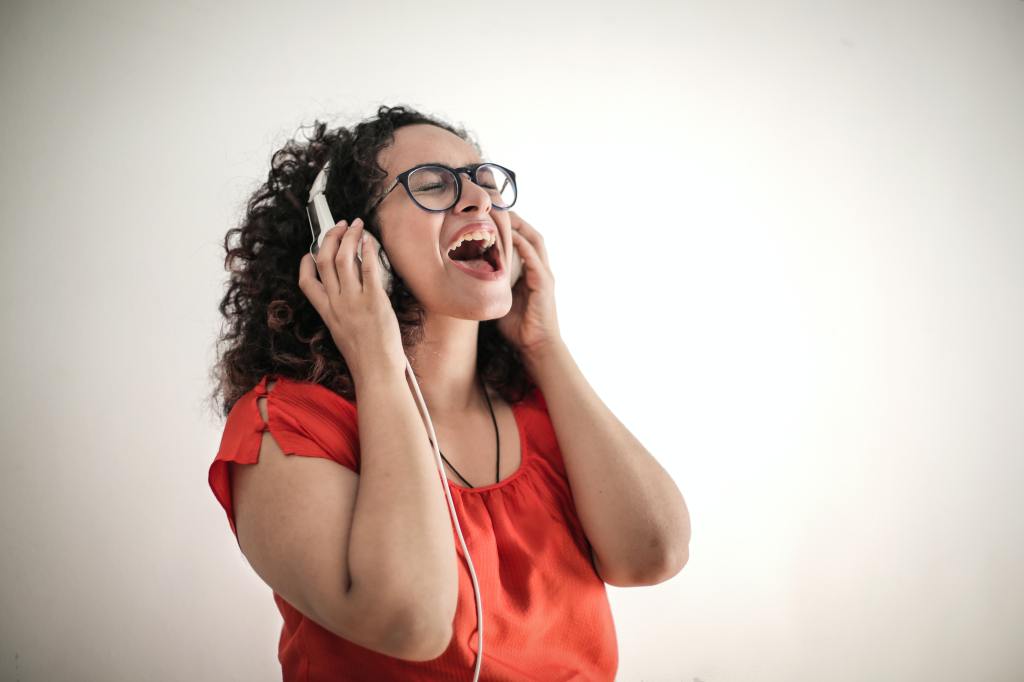 A woman wearing glasses sings passionately with her eyes closed as she holds onto the headphones she's wearing.