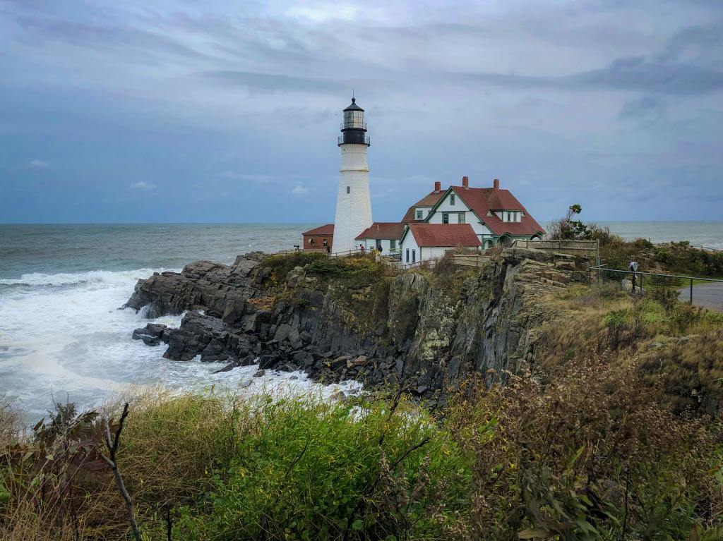 View of a mountain side next to the ocean in Maine. On the mountain are buildings, including a lighthouse. 