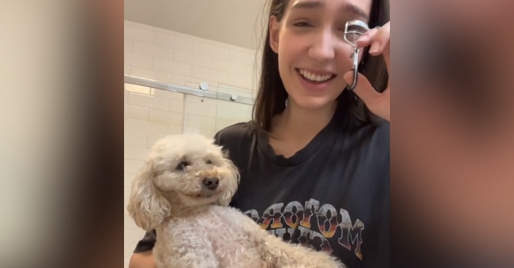 Close up of a woman holding her dog. The dog is smiling as he's cradled in one of her arms. With her free hand, the woman curls her eyelashes and laughs.