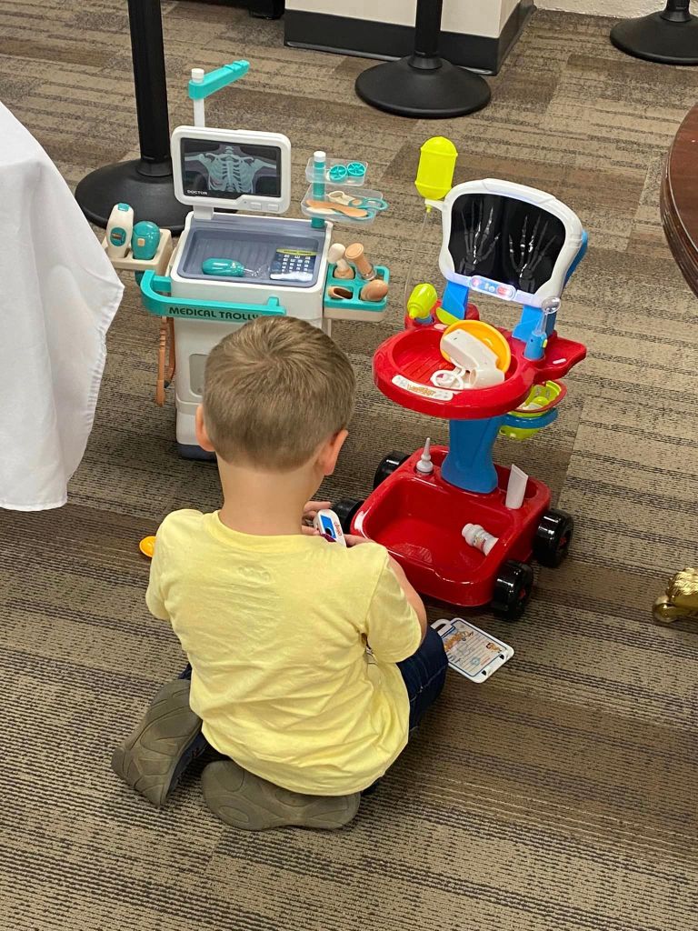 View of a little boy from behind as he sits on the floor, playing with medical equipment toys, including an x-ray machine.