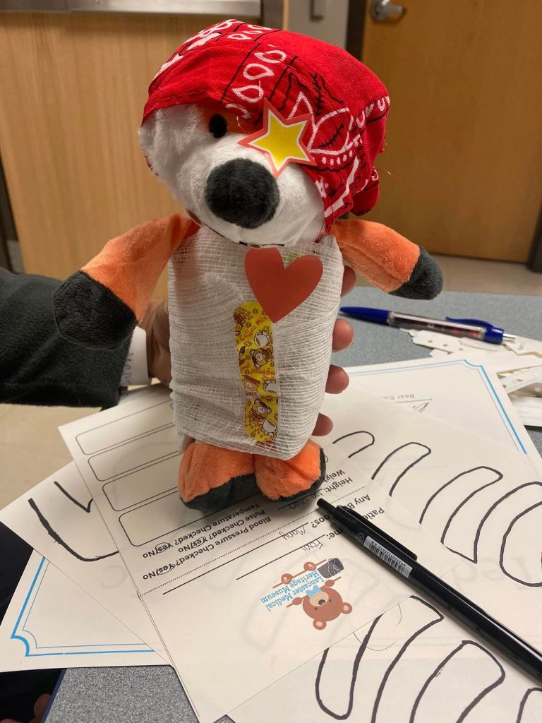 Close up of someone holding up a stuffed fox who has been cared for at the Teddy Bear clinic. The stuffed animal is wearing a bandana, a star sticker as an eye patch, is wrapped in gauze, has a Band-Aid over the gauze, and a heart sticker over where their heart would be.