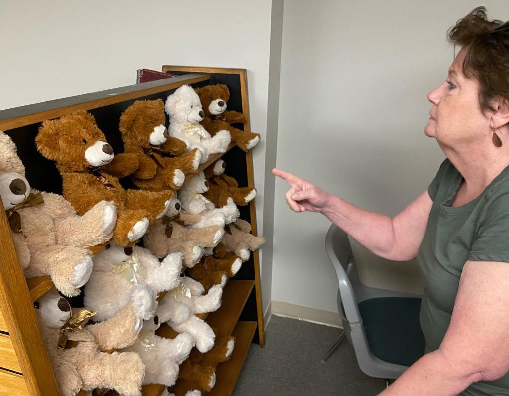 A woman sits facing a shelf full of teddy bears. She's pointing at them as if she's talking to them.