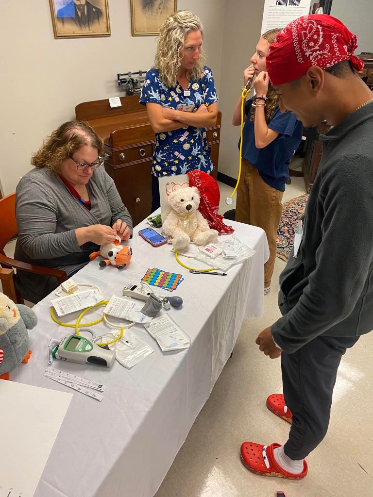 A young person stands in front of a table at the Teddy Bear Clinic at the Lancaster Medical Heritage Museum. On the table are various medical equipment. A woman sitting at the table is treating a stuffed fox. Two medical professionals stand next to the table, talking.