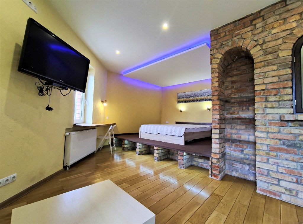 A bedroom with a weird layout. There's a brick wall with built-in shelves with an alcove for the bed that's on a risen platform. The tv is on the opposite wall of the bed and is at an angle where it doesn't seem it can be viewed from the bed.