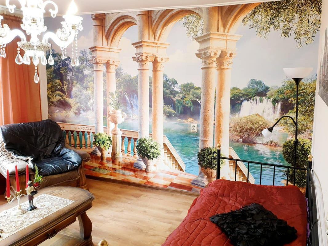 Room with a wall that's entirely covered by a detailed mural. There are giant pillars and a staircase that leads to beautiful water, trees, and a waterfall in the distance.