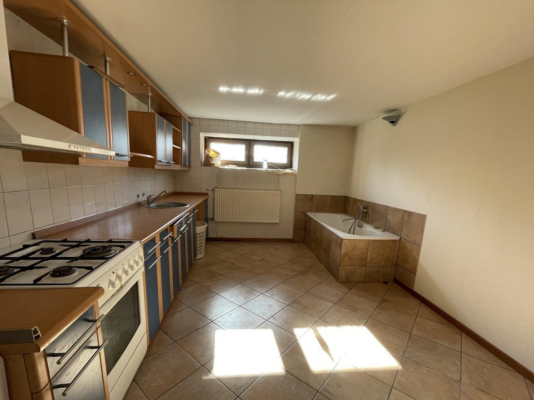 A kitchen where all of the countertops and cabinets are on the left side against the wall. There are also drawers, a stove, and a sink. On the right side is almost an entirely empty wall except for a tub in the very back corner. There's only a few feet between the kitchen side and the bathtub side and nothing dividing the two spaces.