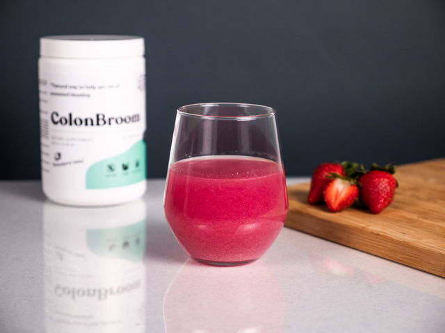 Close up of a clear glass of a red drink sitting on a kitchen counter. Next to it are a few strawberries. On the other side is ColonBroom.