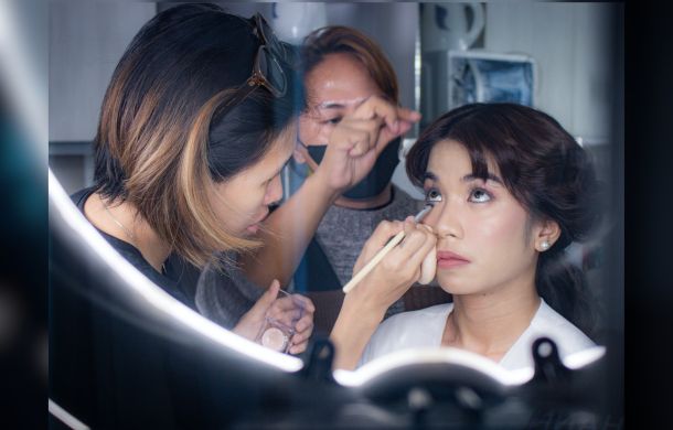 Image is a stock photo showing a bride having her hair and makeup done by two ladies.