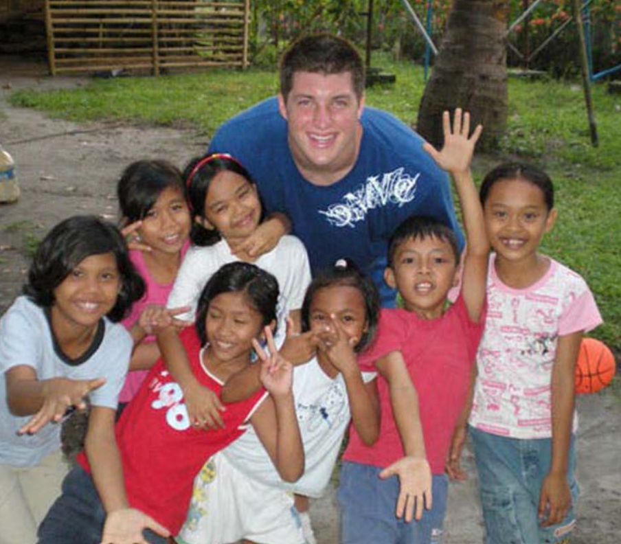 15-year-old Tim Tebow smiling as he poses for a photo with seven little kids.