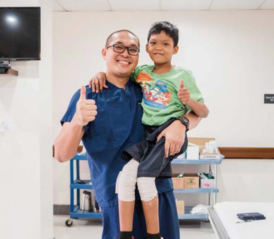 A medical professional smiles and gives a thumbs up as he holds a little boy in his arms, who is also smiling and giving a thumbs up. The little boy has wrappings on his legs, indicating he may have had a procedure.