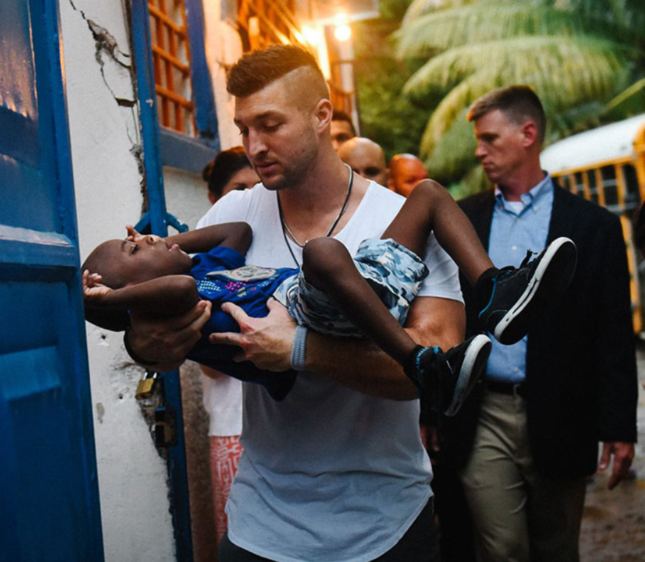 Tim Tebow looks down at the child he's carrying in his arms. A group of people follow behind them as they walk through what appears to be a village.