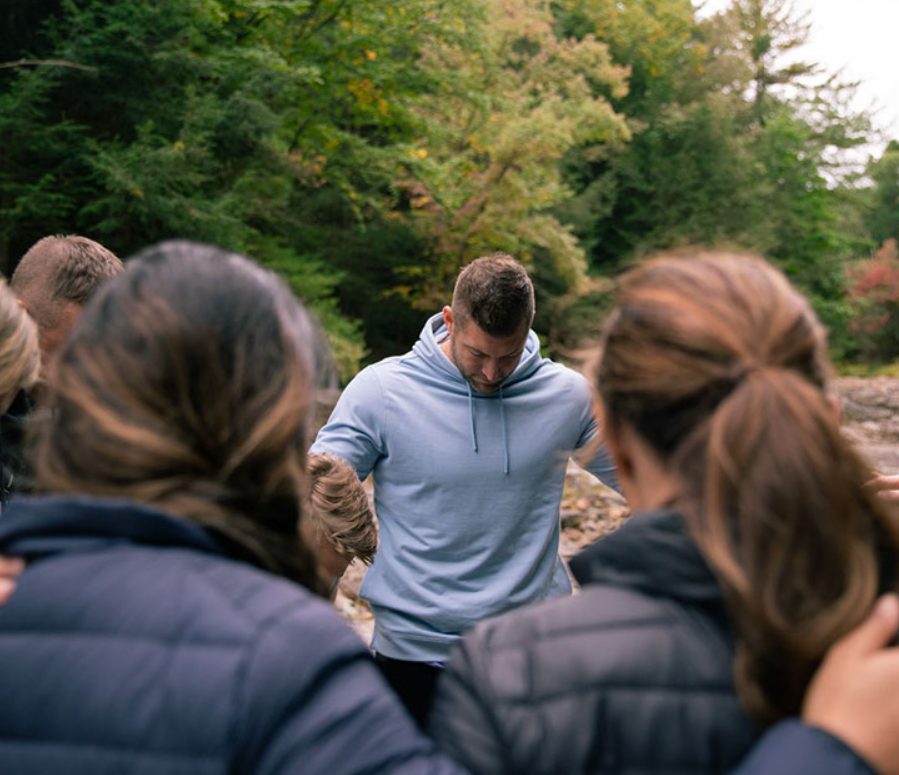 Time Tebow, along with others, stand in a circle, arms around one another, as the bow their heads in prayer.