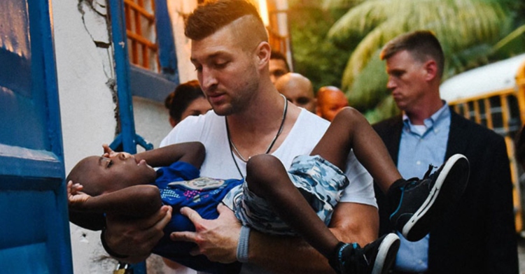 Tim Tebow looks down at the child he's carrying in his arms. A group of people follow behind them as they walk through what appears to be a village.