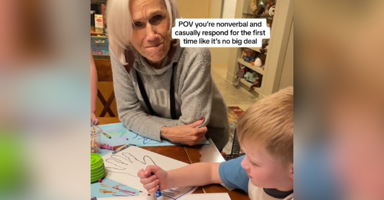 Grandma sits at a table with her grandson who is drawing. She's looking up at the camera, an emotional look on her face.