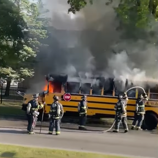 Five firefighters work to put out the flames in the school bus Imunek Williams was driving. Flames can be seen in the drivers seat and lots of thick, black and grey smoke is coming out of all the windows.