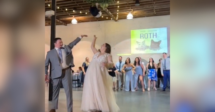 A bride and groom throw a cat plushie for the "cat toss" at their wedding. Guests stand at a distance behind them, getting ready to catch the cat.