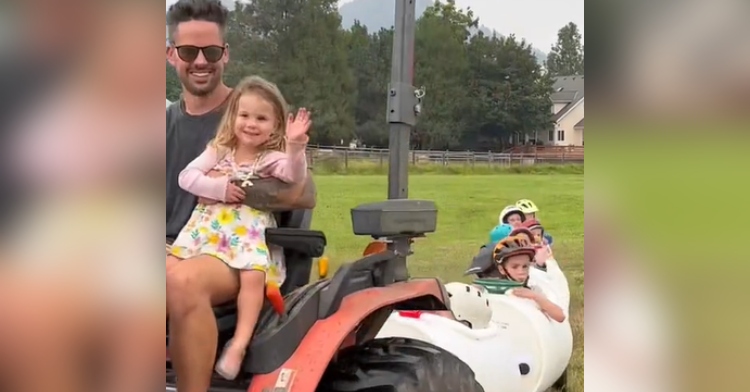 Dad smiles as he drives a tractor. His daughter is sitting in his lap, waving and smiling. The tractor is pulling a line of homemade barrel seats filled with kids.