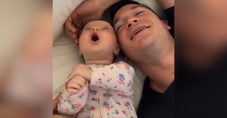 A 3-month-old baby and her dad lay on their backs on a bed, mouths open as they sing.