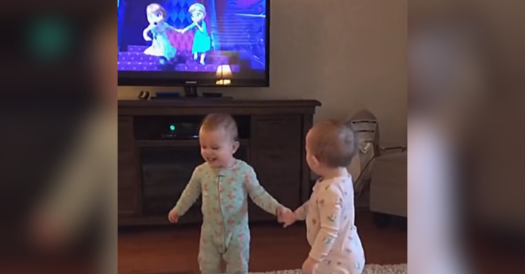 Two twin babies hold hands, mimicking Anna and Elsa from Disney's "Frozen" which can be seen on the TV in front of them.