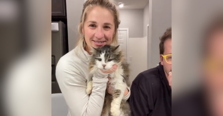 A woman smiles as she holds up her 13-year-old cat named Kitty. The cat is fluffy and mainly white on the front with grey on her head and back.