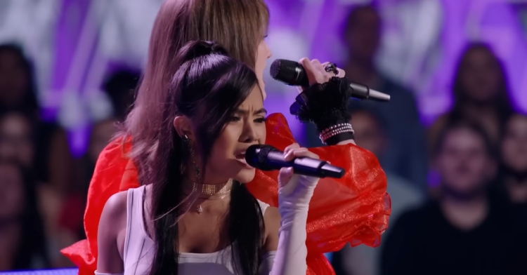 Kaylee Shimizu and Elizabeth Evans sing on "The Voice" stage while standing back to back. The view shows us Kaylee from the front and we see Elizabeth from the back with a side-view of her face.