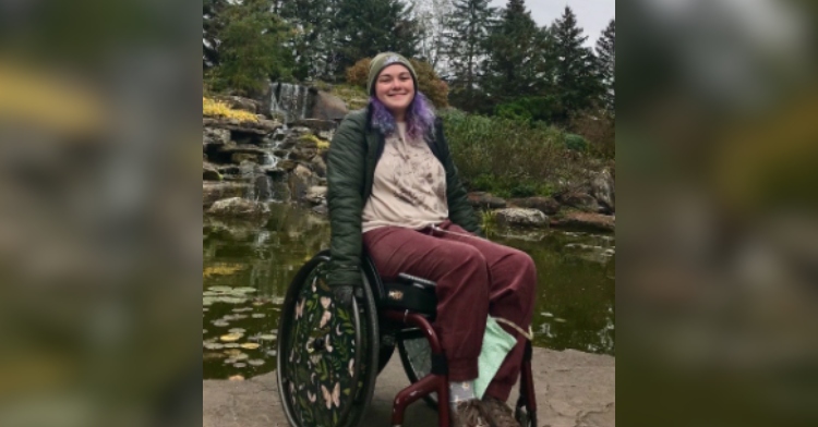 Kyla Greenhunt smiles as she sits in ehr wheelchair. Her hair is now purple. She's near water with a fountain and lots of trees. She looks happy and healthy.