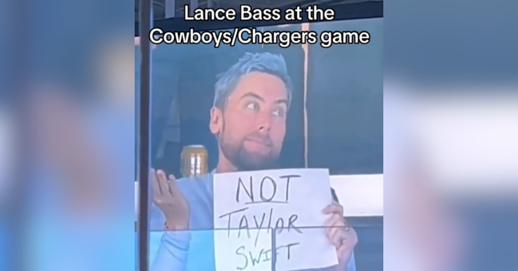Lance Bass shrugs as he holds up a white piece of paper that reads "Not Taylor Swift." He's at a Cowboys/Chargers game.