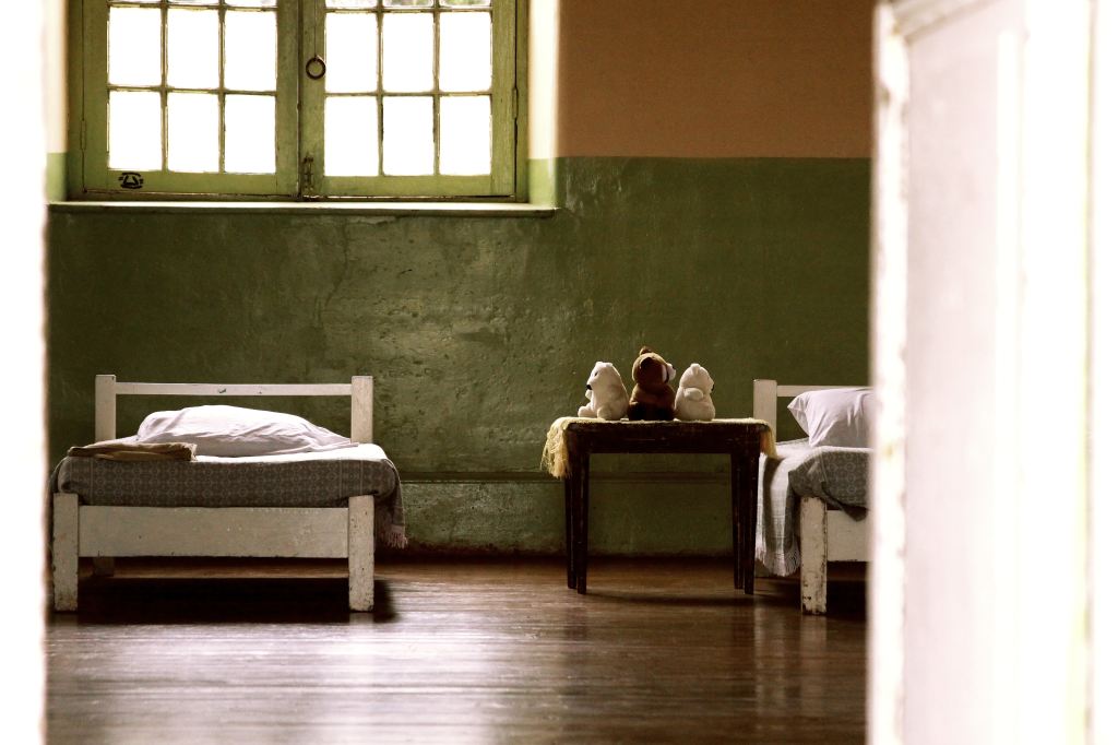 View inside of an orphanage bedroom. The building looks old. Two beds can be seen near a table with three stuffed animals.