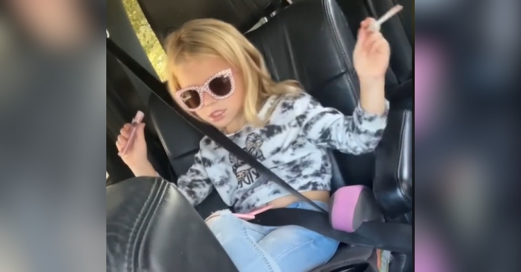 A little girl sits in the backseat of a car. She's wearing sparkly sunglasses and dances as she holds an open lip gloss in her hands.