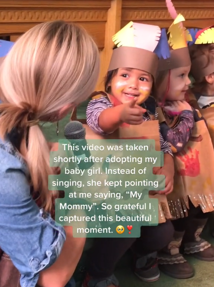 A little girl smiles as she points to her Mom while looking at her teacher. She's standing with other kids who are singing. Text on the image reads: "This video was taken shortly after adopting my baby girl. Instead of singing, she kept pointing at me saying, 'My Mommy.' So grateful I captured this beautiful moment."
