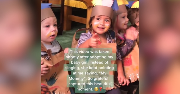 Kids are lined up for a school Thanksgiving performance. One girl smiles as she points at her Mom who is the one capturing this moment on camera. Text on the image reads: "This video was taken shortly after adopting my baby girl. Instead of singing, she kept pointing at me saying, 'My Mommy.' So grateful I captured this beautiful moment."