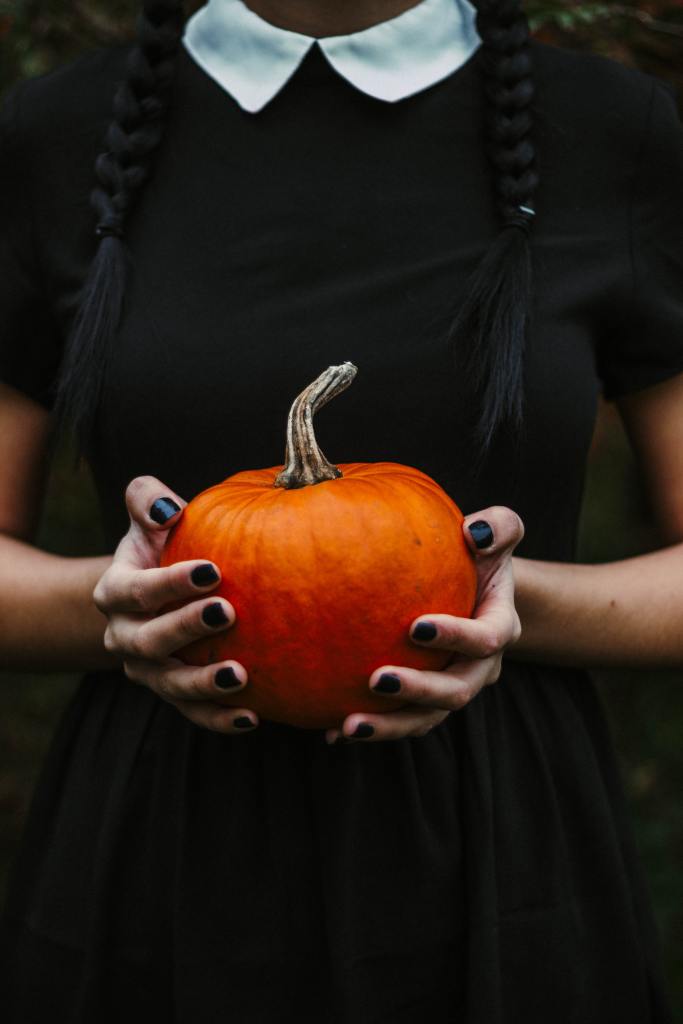 View of a woman from the neck down. She's wearing a black dress with a white collar just like Wednesday Addams. She's holding up a small pumpkin which shows off her black nails.