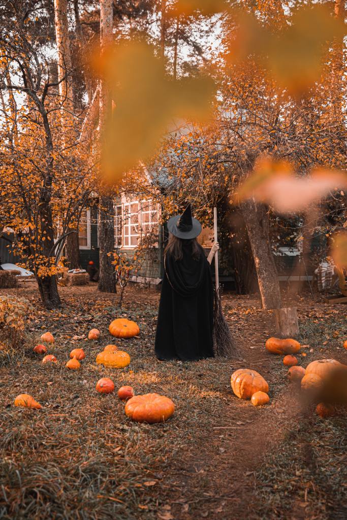 View of a woman from behind who is dressed as a witch with a hat and a broom. She's standing outside and around trees with fall leaves and pumpkins scattered about.