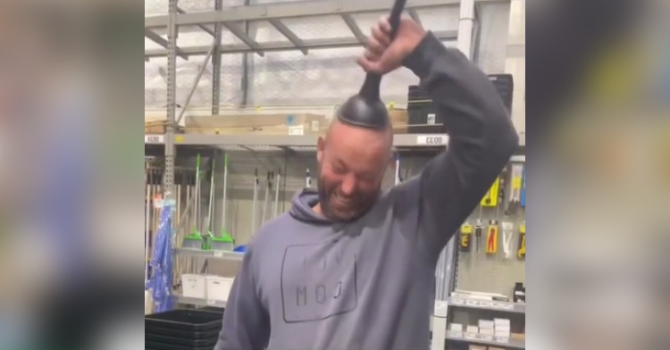 A bald man closes his eyes and concentrates as he tries to pull a plunger off of his head while standing in a store.