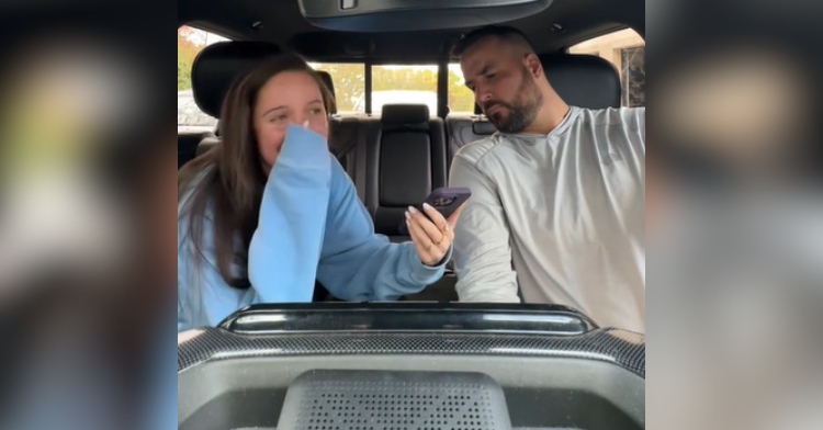 Man and woman sitting in a car. The woman is showing the man something on her phone. With her free hand, the woman covers her mouth, looking nervous. The man looks confused as he watches the woman's phone.