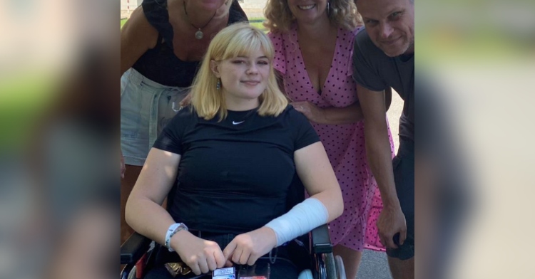 Romy smiles softly from her wheelchair. She is posing for a photo outside with three family members who are smiling. Text on the image reads: "My family thought this was the end stage of my life. They thought I wouldn't be able to live without my legs."