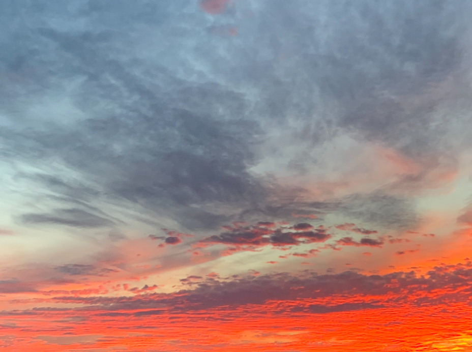 A beautiful sunset taken by Romy. The sky at the top of the photo is a grey/blue color with thin clouds. The sky at the bottom half of the photo is a deep, rich orange/red with more clouds.