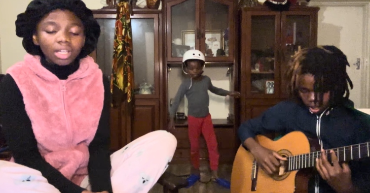 Two siblings sit near the camera, one singing with their eyes closed while the other sings and plays the guitar. Behind them is a third sibling who dances as he wears a helmet and swimming flippers.