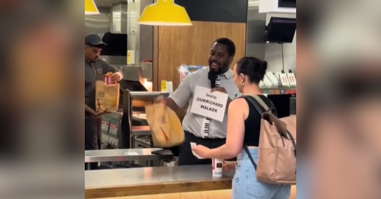 A McDonald's worker smiles as he hands a woman her order. He is also singing into a mic and has a sign that displays his social media username.